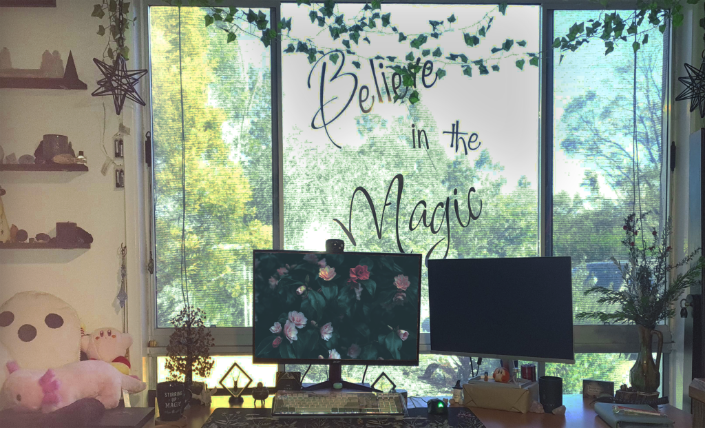 My home office setup by a window with the phrase "Believe in the Magic" on it. The desk has a monitor displaying flowers, another screen, a keyboard, mouse, and various plants. Shelves with decorations and plush toys are visible to the left. The outdoor scenery is visible.