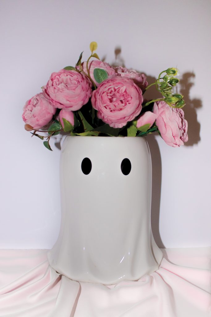 A white ceramic vase shaped like a ghost with black eyes holds a bouquet of pink peonies and green foliage. The vase is set against a plain white background with a light pink draped cloth partially visible at the bottom.