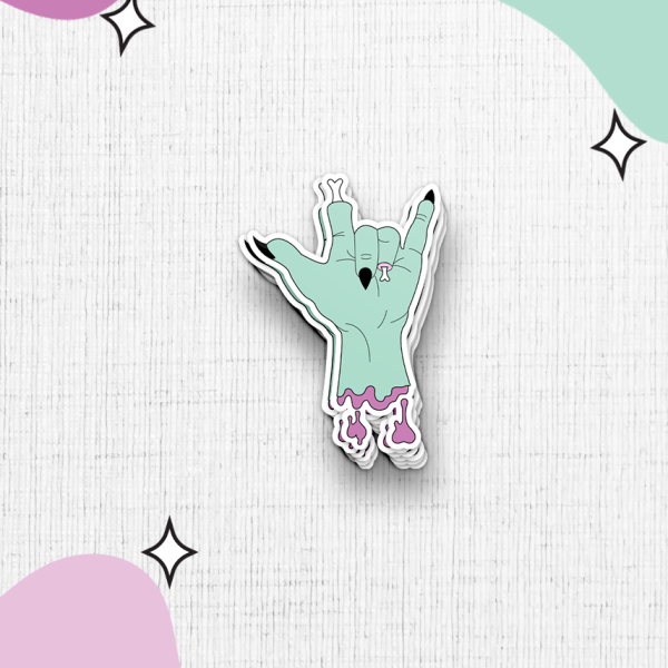 The zombie hand sticker features a greenish hand making the "rock on" gesture with torn flesh near the wrist, set against a textured white background. The image is accented with abstract shapes in pink and mint green and black star-like sparkles.