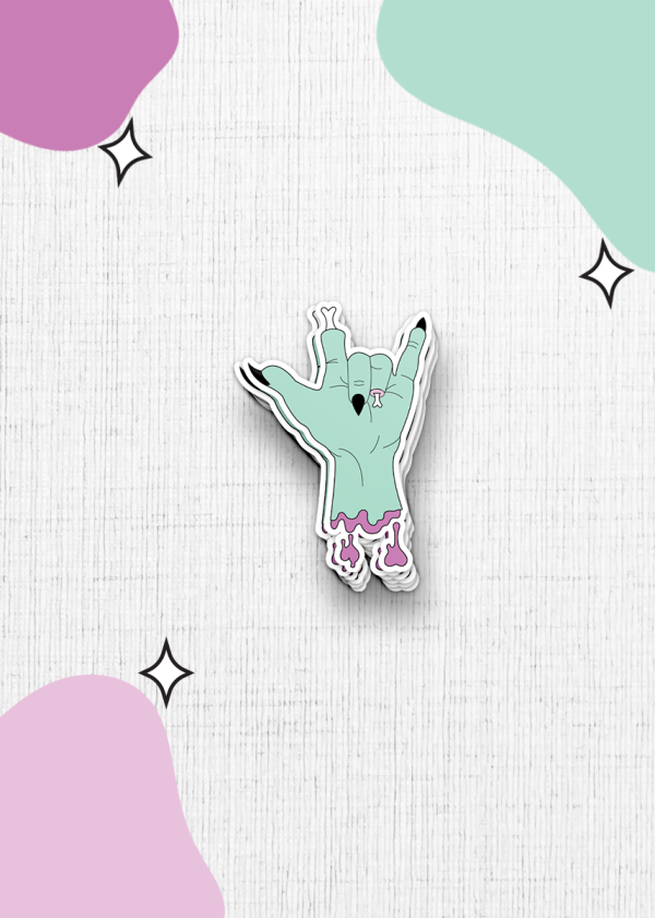 The zombie hand sticker features a greenish hand making the "rock on" gesture with torn flesh near the wrist, set against a textured white background. The image is accented with abstract shapes in pink and mint green and black star-like sparkles.