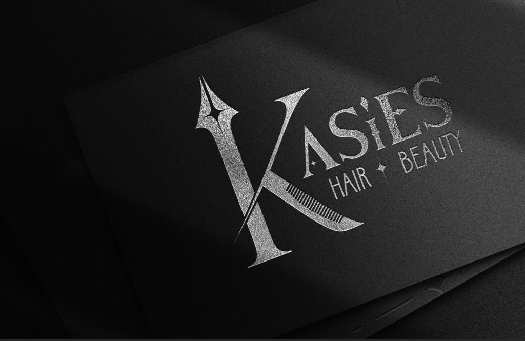 A black card with silver metallic text that reads "Kasie's Hair + Beauty" with a large stylized letter "K" on the left side. The background is shadowy and dark, allowing the text to stand out prominently.