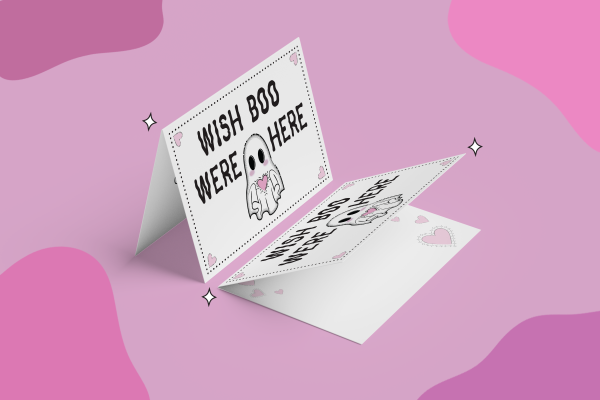 A folded greeting card is displayed against a pink backdrop. The front of the card features a cute ghost illustration with the text "WISH BOO WERE HERE" in a bold, black font. Small hearts and twinkling star-like shapes decorate the card, adding a whimsical touch.