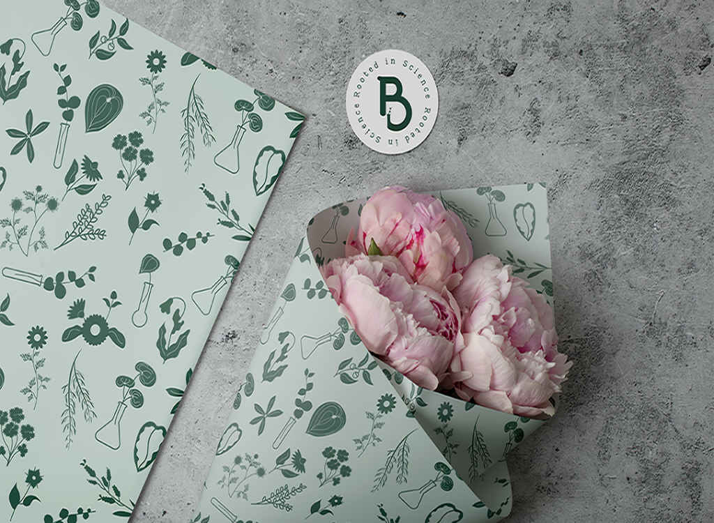 A bouquet of light pink peonies wrapped in light green paper adorned with botanical illustrations, placed on a gray, textured surface. Near the bouquet is a round card with a "B" logo and text around it. Another sheet of the same wrapping paper is spread out beside the bouquet.