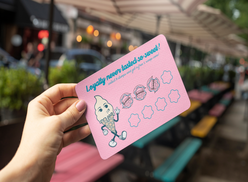 A hand holds a pink loyalty card for an ice cream shop. The card features a cartoon ice cream cone character and the text "Loyalty never tasted so sweet!" Beneath it are spaces for stamps, with three already stamped. Outdoor seating and a street are visible in the background.